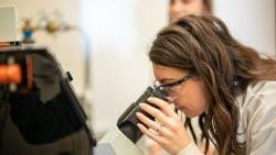 Woman wearing protective goggles looking through a microscope.